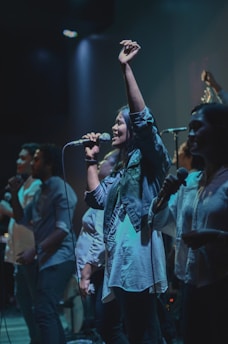 A group of people is performing on stage with microphones, captured under dim, dramatic lighting. The main focus is on a woman singing passionately with her arm raised.