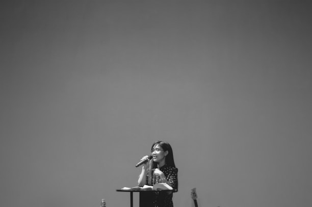 A woman is standing behind a podium, speaking into a microphone. She has a book or papers on the podium and appears to be giving a speech or presentation. The background is plain and minimalistic.