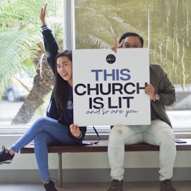 Two people are sitting on a bench inside a building, holding a large sign that reads 'THIS CHURCH IS LIT and so are you.' One person has their arm raised and is smiling, while the other gives a peace sign. There are large windows and plants visible in the background.
