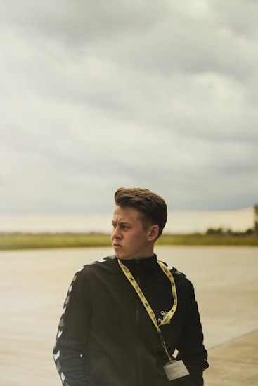 A person with short brown hair is standing outdoors on an overcast day. They are wearing a black jacket with white details on the sleeves and a yellow lanyard around their neck that holds an ID badge. The background shows a flat, open area with grass and a cloudy sky.