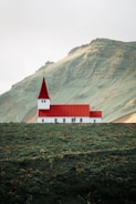 A small church with a red roof and white walls is situated on a lush green hillside. The background features a large mountain with an earthy texture. The scene appears tranquil and isolated, with minimal human presence.