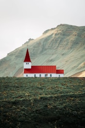 A small church with a red roof and white walls is situated on a lush green hillside. The background features a large mountain with an earthy texture. The scene appears tranquil and isolated, with minimal human presence.