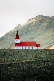A small church with a red roof and white walls is situated on a lush green hillside. The background features a large mountain with an earthy texture. The scene appears tranquil and isolated, with minimal human presence.