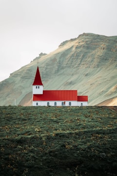 A small church with a red roof and white walls is situated on a lush green hillside. The background features a large mountain with an earthy texture. The scene appears tranquil and isolated, with minimal human presence.