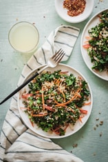 A fresh salad with kale, quinoa, carrots, and other greens is served on a white plate. A fork is placed on a striped cloth next to the plate. A small dish with red pepper flakes and a glass of light-colored drink are nearby.