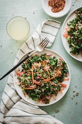 A fresh salad with kale, quinoa, carrots, and other greens is served on a white plate. A fork is placed on a striped cloth next to the plate. A small dish with red pepper flakes and a glass of light-colored drink are nearby.