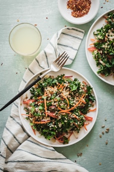 A fresh salad with kale, quinoa, carrots, and other greens is served on a white plate. A fork is placed on a striped cloth next to the plate. A small dish with red pepper flakes and a glass of light-colored drink are nearby.