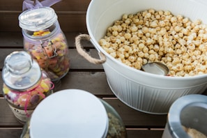 A large white bowl filled with popcorn is placed on a wooden surface. Next to it are glass jars, some with colorful candy, including gummy worms and other sweets. One jar is adorned with a pink ribbon.