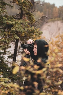 A person wearing a black hoodie is taking a photograph with a camera in a forested area. The scene is surrounded by trees and foliage, showcasing a natural and serene environment.
