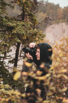 A person wearing a black hoodie is taking a photograph with a camera in a forested area. The scene is surrounded by trees and foliage, showcasing a natural and serene environment.