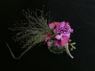 A floral arrangement featuring vibrant pink and purple flowers, adorned with delicate, wispy green foliage. The flowers are intricately detailed, set against a dark, contrasting background that accentuates their colors.