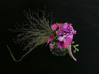 A floral arrangement featuring vibrant pink and purple flowers, adorned with delicate, wispy green foliage. The flowers are intricately detailed, set against a dark, contrasting background that accentuates their colors.
