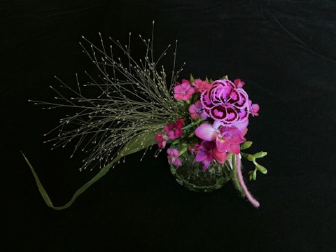 A floral arrangement featuring vibrant pink and purple flowers, adorned with delicate, wispy green foliage. The flowers are intricately detailed, set against a dark, contrasting background that accentuates their colors.