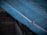 A close-up view of a weathered, blue-tinted metal surface with a visible bolt or fastener. The metal appears to be part of a roofing structure and has signs of wear, with some rust visible around the bolt.