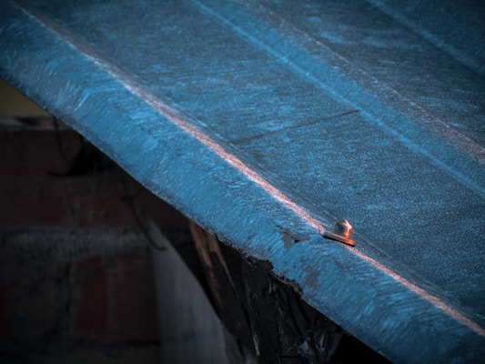A close-up view of a weathered, blue-tinted metal surface with a visible bolt or fastener. The metal appears to be part of a roofing structure and has signs of wear, with some rust visible around the bolt.
