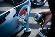 A close-up of a skateboarding scene with a skateboard deck featuring artistic graphics and a hand holding a tool for assembling a wheel. The surface is detailed with colorful designs and a wheel is decorated with intricate patterns. A person's sneaker is partially visible at the bottom of the image.