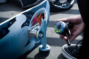 A close-up of a skateboarding scene with a skateboard deck featuring artistic graphics and a hand holding a tool for assembling a wheel. The surface is detailed with colorful designs and a wheel is decorated with intricate patterns. A person's sneaker is partially visible at the bottom of the image.