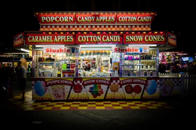 A brightly lit concession stand at a carnival offers popcorn, candy apples, cotton candy, caramel apples, and snow cones. The front of the stand features vibrant signage and images of the treats available. Inside, the stand is neatly stocked with various colorful products and displays of beverages with 'Slushie' signs.