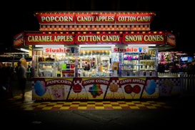 A brightly lit concession stand at a carnival offers popcorn, candy apples, cotton candy, caramel apples, and snow cones. The front of the stand features vibrant signage and images of the treats available. Inside, the stand is neatly stocked with various colorful products and displays of beverages with 'Slushie' signs.