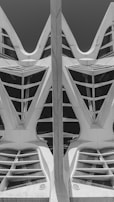 A black and white photograph featuring an architectural structure with geometric patterns. The design includes a combination of sharp and curved edges, creating a symmetrical and futuristic appearance. The structure has triangular and semi-circular openings, with a central vertical element dividing the scene.