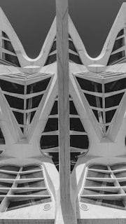 A black and white photograph featuring an architectural structure with geometric patterns. The design includes a combination of sharp and curved edges, creating a symmetrical and futuristic appearance. The structure has triangular and semi-circular openings, with a central vertical element dividing the scene.