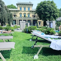 A luxurious villa serves as the backdrop to a lush garden area with sun loungers scattered on a well-manicured lawn. The building is a grand, multi-story structure with multiple windows and balconies. Tall, mature trees flank the garden, providing a sense of privacy and tranquility. A few people are visible, some relaxing on the loungers while others enjoy the nearby pool. There's a sense of summer leisure and relaxation captured in the scene.