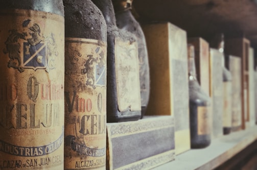 Close-up photo of an aged wine bottle with a detailed vintage label and slight dust, resting gently on a rustic wooden shelf in a dimly lit cellar.
