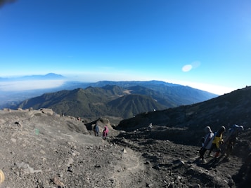 A rugged mountainous landscape with a clear blue sky, overlooking a range of green and rocky terrains. Several hikers with backpacks are making their way up a steep, rocky trail. The distant horizon reveals a softer mist, suggesting the early morning or just after sunrise.