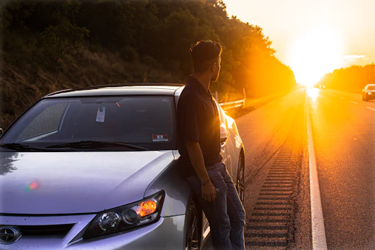Smiling roadside assistance technician helping a relieved driver by a modern car at sunset