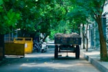 A narrow road shaded by lush green trees on either side. A vehicle resembling a small truck or cart is filled with sand and parked on the right side of the road. Several scooters are parked nearby along the pavement. There is a large yellow bin on the left side.