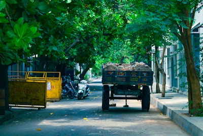 A narrow road shaded by lush green trees on either side. A vehicle resembling a small truck or cart is filled with sand and parked on the right side of the road. Several scooters are parked nearby along the pavement. There is a large yellow bin on the left side.