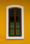 A wooden window with vertical bars is set against a vibrant yellow wall. The window frame is brown and the glass panes are dark, creating contrast with the surrounding white border.