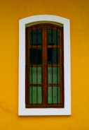 A wooden window with vertical bars is set against a vibrant yellow wall. The window frame is brown and the glass panes are dark, creating contrast with the surrounding white border.