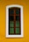 A wooden window with vertical bars is set against a vibrant yellow wall. The window frame is brown and the glass panes are dark, creating contrast with the surrounding white border.