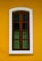 A wooden window with vertical bars is set against a vibrant yellow wall. The window frame is brown and the glass panes are dark, creating contrast with the surrounding white border.