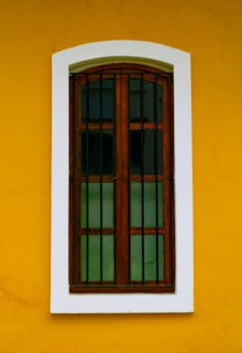 A wooden window with vertical bars is set against a vibrant yellow wall. The window frame is brown and the glass panes are dark, creating contrast with the surrounding white border.