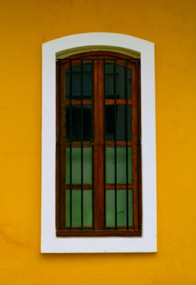 A wooden window with vertical bars is set against a vibrant yellow wall. The window frame is brown and the glass panes are dark, creating contrast with the surrounding white border.