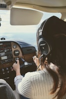 A person with long hair wearing a headset is piloting a small aircraft. The view is from behind the pilot, showing their hands on the controls and the cockpit dashboard with various instruments and panels. The scene suggests that the aircraft is in-flight with a view of the sky and horizon through the windshield.