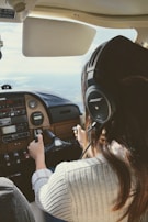 A person with long hair wearing a headset is piloting a small aircraft. The view is from behind the pilot, showing their hands on the controls and the cockpit dashboard with various instruments and panels. The scene suggests that the aircraft is in-flight with a view of the sky and horizon through the windshield.