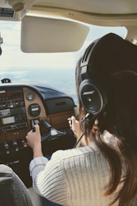 A person with long hair wearing a headset is piloting a small aircraft. The view is from behind the pilot, showing their hands on the controls and the cockpit dashboard with various instruments and panels. The scene suggests that the aircraft is in-flight with a view of the sky and horizon through the windshield.
