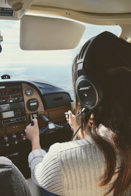 A person with long hair wearing a headset is piloting a small aircraft. The view is from behind the pilot, showing their hands on the controls and the cockpit dashboard with various instruments and panels. The scene suggests that the aircraft is in-flight with a view of the sky and horizon through the windshield.