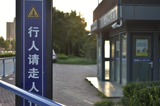 A pedestrian sign with Chinese characters stands tall beside a pathway. The background includes a small building with glass windows and a sign that reads 'Traffic Police' in both English and Chinese. Greenery and a partially visible car are present, and the lighting suggests late afternoon or early evening.