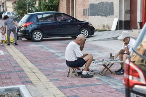 Two elderly men are playing a board game on a sidewalk, sitting on small stools. They are deeply focused on the game, with one holding his head in his hand. A black car is parked nearby. In the background, a woman and a child are seen walking away, with the child holding a yellow object.