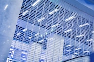A metal gate with a grid pattern fills the foreground, with bright fluorescent lighting and ceiling structures visible through the gaps. The scene is dominated by cool tones, creating a modern and industrial atmosphere.