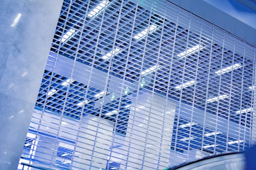 A metal gate with a grid pattern fills the foreground, with bright fluorescent lighting and ceiling structures visible through the gaps. The scene is dominated by cool tones, creating a modern and industrial atmosphere.