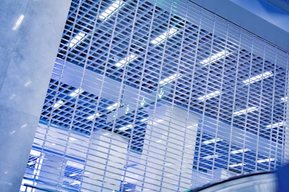A metal gate with a grid pattern fills the foreground, with bright fluorescent lighting and ceiling structures visible through the gaps. The scene is dominated by cool tones, creating a modern and industrial atmosphere.