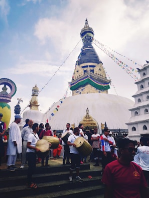 A large stupa with iconic eyes and colorful prayer flags, surrounded by a group of people, some playing traditional drums. The sky is partly cloudy, creating a vibrant yet serene atmosphere.