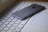 A smartphone is placed face down on the keyboard of a laptop, creating a modern tech workspace setting. The focus is on the smartphone, and the keyboard keys are visible around it.