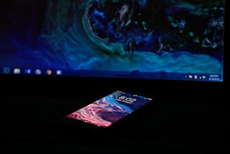 A smartphone with a colorful, abstract lock screen is lying on a surface in front of a laptop. The laptop's screen displays a similar abstract pattern with blue and green hues, and the desktop icons are dimly visible. The setting is dark, implying a low-light environment.