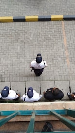 A group of security personnel wearing uniforms and caps standing on a tiled pavement, viewed from above. The tiles are organized in a geometric pattern, and there's a black and yellow curb visible at the top of the image.